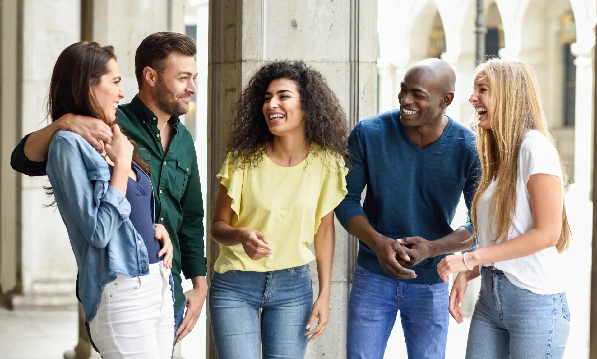 A group of five friends stands together, laughing and chatting, in a historic archway with stone columns and a light background